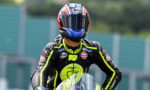 Motorcycle racer in full gear and helmet standing on track, neon-yellow accents and sponsor logos visible (Gulf, Michelin).