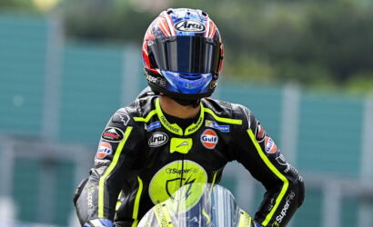 Motorcycle racer in full gear and helmet standing on track, neon-yellow accents and sponsor logos visible (Gulf, Michelin).