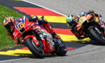 Two professional motorcycle racers lean into a sharp turn on a race track, red and orange bikes closely side by side with yellow-red-black curbing in the background.