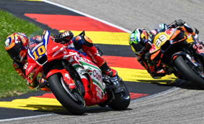 Two professional motorcycle racers lean into a sharp turn on a race track, red and orange bikes closely side by side with yellow-red-black curbing in the background.