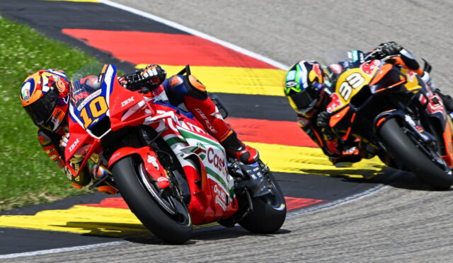 Two professional motorcycle racers lean into a sharp turn on a race track, red and orange bikes closely side by side with yellow-red-black curbing in the background.