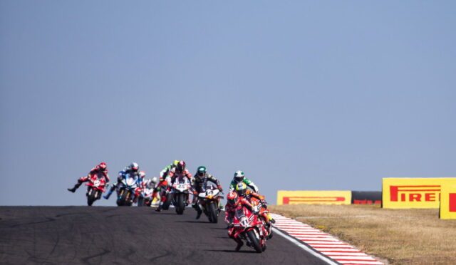 Group of motorcycle racers lean into a track bend on a racetrack with Pirelli barriers in the background.