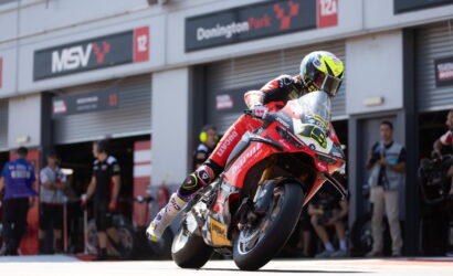 Moto racer leaning sharply into a turn on a red racing bike, pit garages in the background.