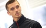 Man in a black Honda jacket with a red zipper, looking to the side in an indoor setting.