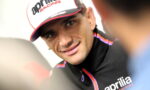 Close-up of a smiling man in an Aprilia Racing cap and black jacket, talking during an interview.