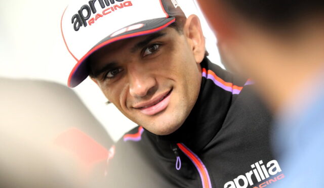 Close-up of a smiling man in an Aprilia Racing cap and black jacket, talking during an interview.