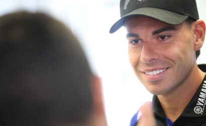 Young man in a black Yamaha cap and lanyard smiles while talking to someone off-camera.