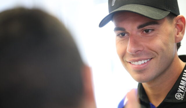 Young man in a black Yamaha cap and lanyard smiles while talking to someone off-camera.