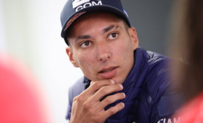 Young man in a navy cap and puffer jacket gazing to his left, with his chin resting on his hand in a thoughtful pose.
