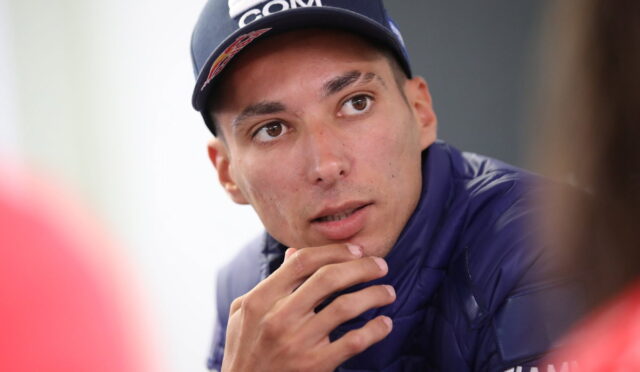 Young man in a navy cap and puffer jacket gazing to his left, with his chin resting on his hand in a thoughtful pose.