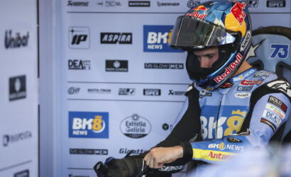 Motorcycle racer in a blue racing suit and Red Bull helmet sits in a pit area against a sponsor backdrop.