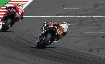 Two professional motorcycle racers lean into a tight corner on a race track, tire tracks curving across the asphalt and red-and-white curb at the inside of the bend visible on the left.