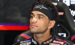 Portrait of a male race car driver with a beige nose strip, wearing a black headband, seated in a racing seat with sponsor logos on the suit