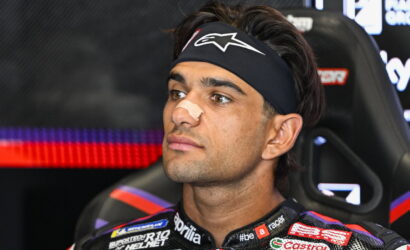 Portrait of a male race car driver with a beige nose strip, wearing a black headband, seated in a racing seat with sponsor logos on the suit