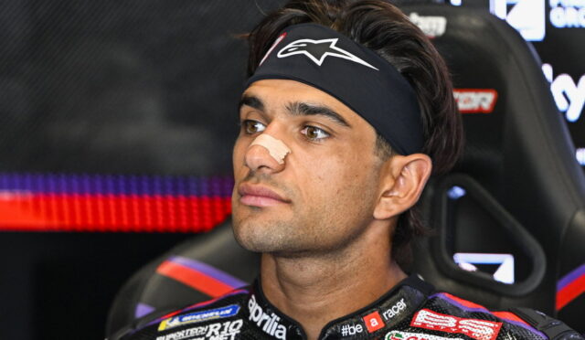 Portrait of a male race car driver with a beige nose strip, wearing a black headband, seated in a racing seat with sponsor logos on the suit