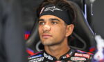 Male motorcycle racer wearing a black headband with a white logo and a beige nose band, seated in a racing chair and gazing ahead.