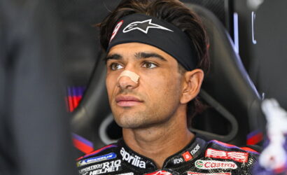 Male motorcycle racer wearing a black headband with a white logo and a beige nose band, seated in a racing chair and gazing ahead.