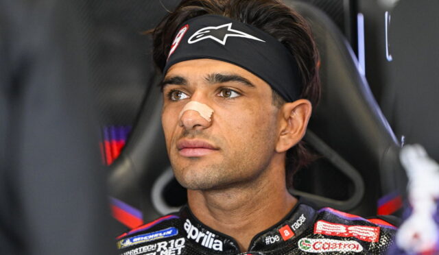Male motorcycle racer wearing a black headband with a white logo and a beige nose band, seated in a racing chair and gazing ahead.