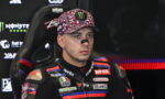 Male motorcycle racer in a sponsor-filled racing suit sits in a chair, wearing a colorful cap with a Monster Energy logo and eye black.