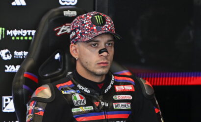 Male motorcycle racer in a sponsor-filled racing suit sits in a chair, wearing a colorful cap with a Monster Energy logo and eye black.