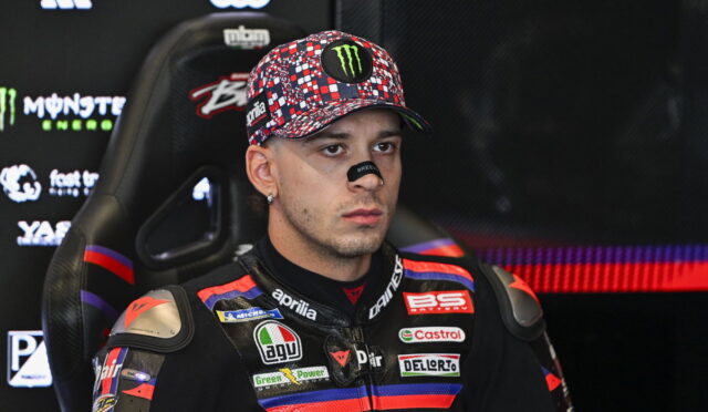 Male motorcycle racer in a sponsor-filled racing suit sits in a chair, wearing a colorful cap with a Monster Energy logo and eye black.