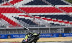 Motorcycle racer in neon yellow gear leans into a turn on a race track, with red, white and blue empty grandstands in the background.