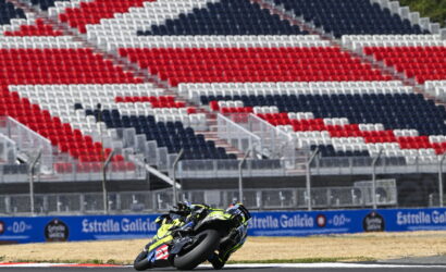 Motorcycle racer in neon yellow gear leans into a turn on a race track, with red, white and blue empty grandstands in the background.