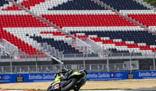 Motorcycle racer in neon yellow gear leans into a turn on a race track, with red, white and blue empty grandstands in the background.