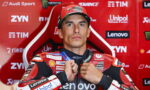 MotoGP rider in a red racing suit sits in a red sponsor backdrop, adjusting his zipper with sponsor logos like Lenovo and Unipol visible on suit.