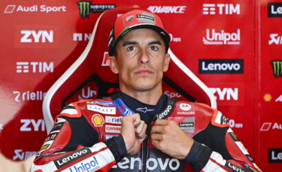 MotoGP rider in a red racing suit sits in a red sponsor backdrop, adjusting his zipper with sponsor logos like Lenovo and Unipol visible on suit.
