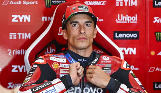 MotoGP rider in a red racing suit sits in a red sponsor backdrop, adjusting his zipper with sponsor logos like Lenovo and Unipol visible on suit.