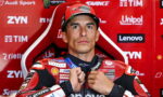 MotoGP rider in a red and black racing suit adjusts his zipper while posing against a red sponsor-filled backdrop.