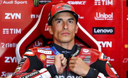 MotoGP rider in a red and black racing suit adjusts his zipper while posing against a red sponsor-filled backdrop.