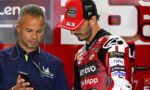 Team member in blue with Michelin logo looks at a smartphone while a MotoGP rider in a red Lenovo cap studies with him in the paddock.