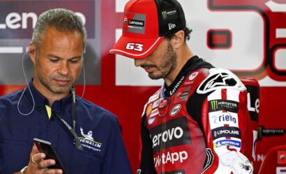 Team member in blue with Michelin logo looks at a smartphone while a MotoGP rider in a red Lenovo cap studies with him in the paddock.