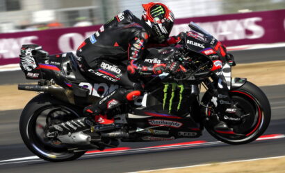 Two professional motorcycle racers lean into a high-speed corner on a sponsor-laden sportbike, sharing the same bike on a track lap.