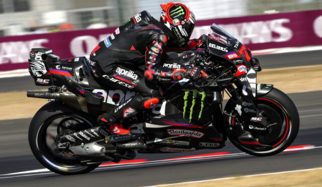 Two professional motorcycle racers lean into a high-speed corner on a sponsor-laden sportbike, sharing the same bike on a track lap.