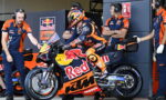 Rider in orange racing gear sits on a Red Bull KTM motorcycle in the pit with team members in blue and orange nearby.