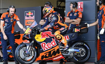 Rider in orange racing gear sits on a Red Bull KTM motorcycle in the pit with team members in blue and orange nearby.