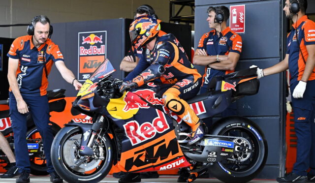 Rider in orange racing gear sits on a Red Bull KTM motorcycle in the pit with team members in blue and orange nearby.