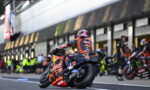 MotoGP rider in orange/blue Red Bull KTM suit leans into a turn on a race bike with #93 livery, helmet pink accents, in the pit lane.