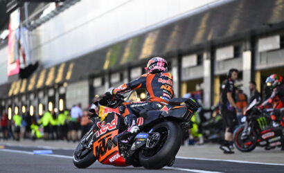 MotoGP rider in orange/blue Red Bull KTM suit leans into a turn on a race bike with #93 livery, helmet pink accents, in the pit lane.