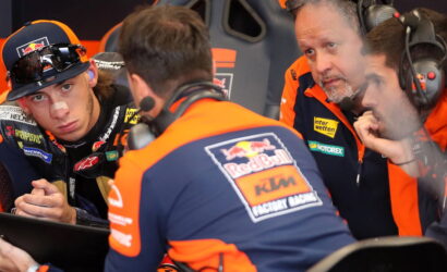 Red Bull KTM Factory Racing crew in orange and blue consult a tablet in the pit, rider with a nose bandage watches nearby.
