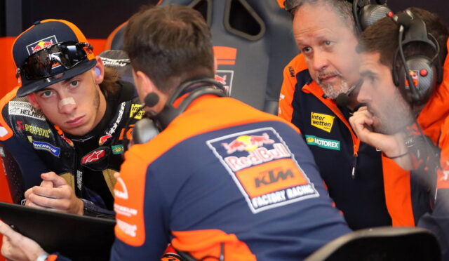 Red Bull KTM Factory Racing crew in orange and blue consult a tablet in the pit, rider with a nose bandage watches nearby.