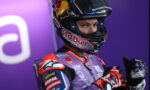 Moto racer in a purple sponsor-laden suit and full-face Red Bull helmet, giving a thumbs-up against a purple background.