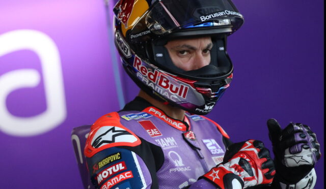 Moto racer in a purple sponsor-laden suit and full-face Red Bull helmet, giving a thumbs-up against a purple background.