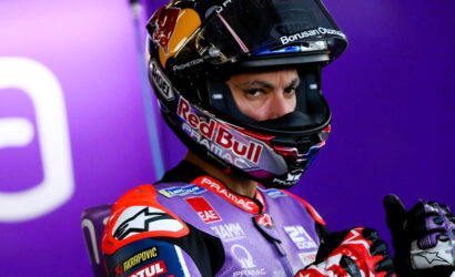 MotoGP rider in a Red Bull helmet and purple racing suit gives a thumbs-up toward the camera against a purple backdrop