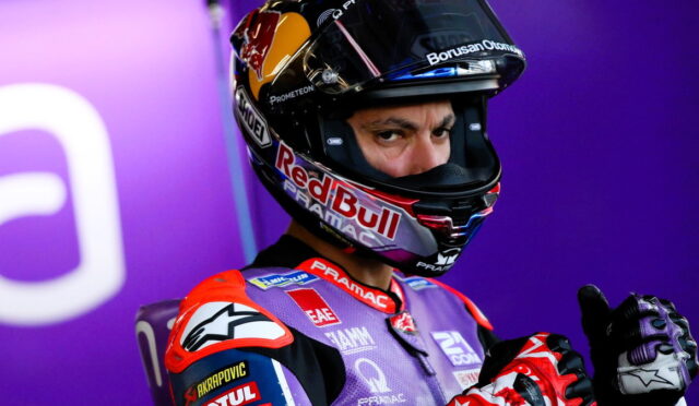 MotoGP rider in a Red Bull helmet and purple racing suit gives a thumbs-up toward the camera against a purple backdrop