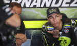 Motocross racer in neon-yellow and black gear sits in a team garage, palm raised as he talks to a teammate beside him.【informative】