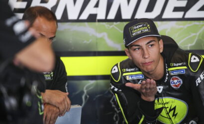 Motocross racer in neon-yellow and black gear sits in a team garage, palm raised as he talks to a teammate beside him.【informative】
